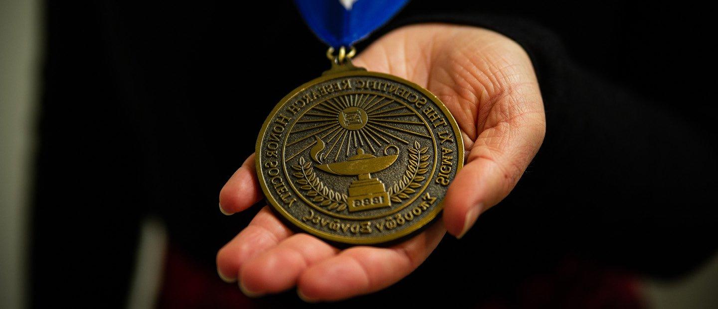 A hand holding a round gold medal. Text on the medal says "Sigma Xi The Scientific Research Honor Society"